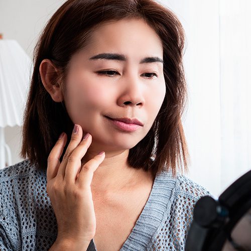 Mulher observa o próprio rosto no espelho, tocando a bochecha com expressão atenta, possivelmente avaliando sinais de paralisia facial.