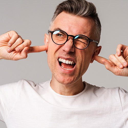Homem com expressão de desconforto, tampando os ouvidos, sugerindo problema auditivo ou dor.