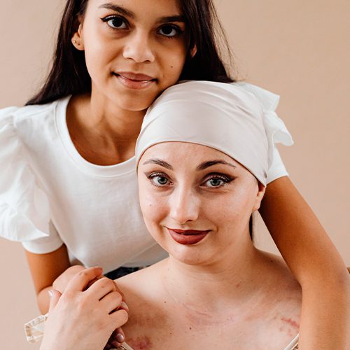 Mulher jovem abraça outra mulher com lenço na cabeça e cicatrizes no colo, simbolizando apoio e força durante tratamento contra o câncer.